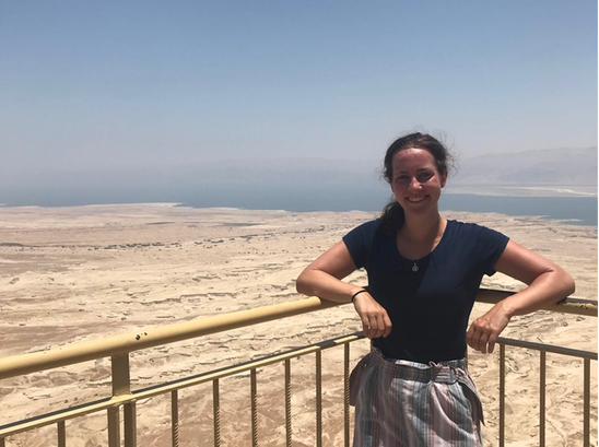 Talia Buchman, an anthropology major, posing in Israel on a railing. She is wearing a blue top, shorts, and is smiling. She has brown hair and blue eyes and is skinny. The terrain behind her is a dusty desert and it mets the blue muddled sky.