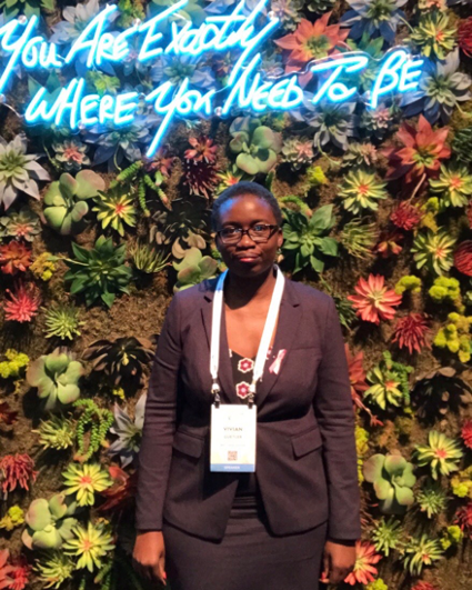 Vivian Guetler, black woman phd candidate at WVU recipient of Outstanding Dissertation Award standing in black suit in front of a greenery plant background and a blue electric sign that says "you are exactly where you need to be"