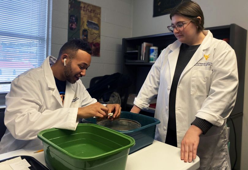 Bryan Hill, left, and Kelsey Gill, right, are wearing white lab coats while cleaning an artifact in the WVU Archaeology Lab.