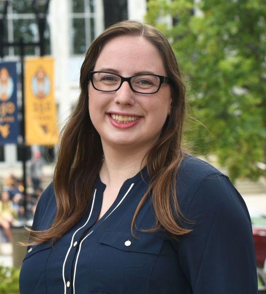 Brittany Kowlaski, Graduate Research Assistant. White woman with black glasses and mid length brown hair smiling at the camera wearing a blue button up with white seams in front of the mountainlair with two wvu blue and gold flags and a tree