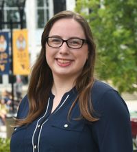 Brittany Kolwaski is in a blue and white button up shirt she smiles while wearing square rectangled glasses in front of a tree and a 150 years of WVU flags in front of a white and gray mountainlair window