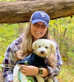 Leah Oldham, PhD student WVU holding white dog. she is white with blonde hair, wearing a blue cap with a flying WV on it. She is located in a forest with a tree above her head